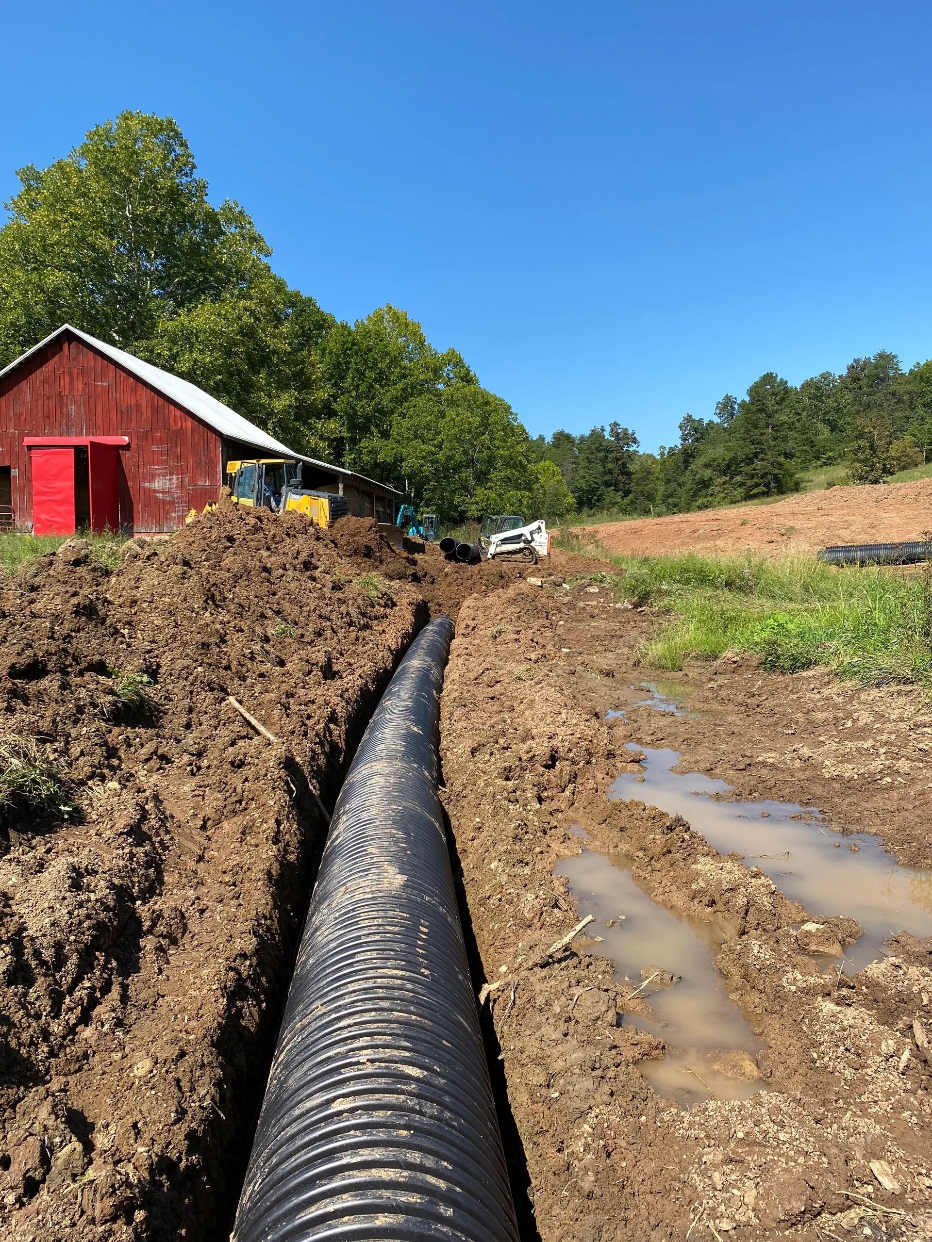 Long drainage pipe in a trench next to a red barn.